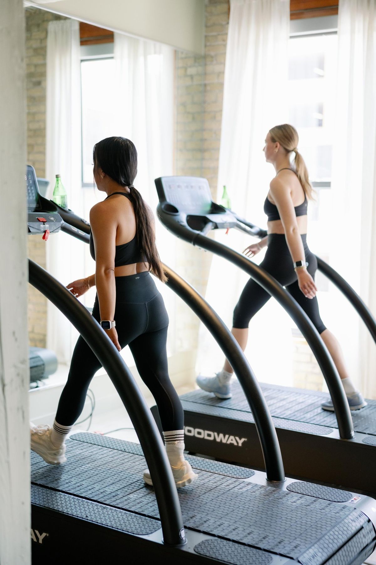 Two members running on Woodway treadmills beside the studio's brick-framed window
